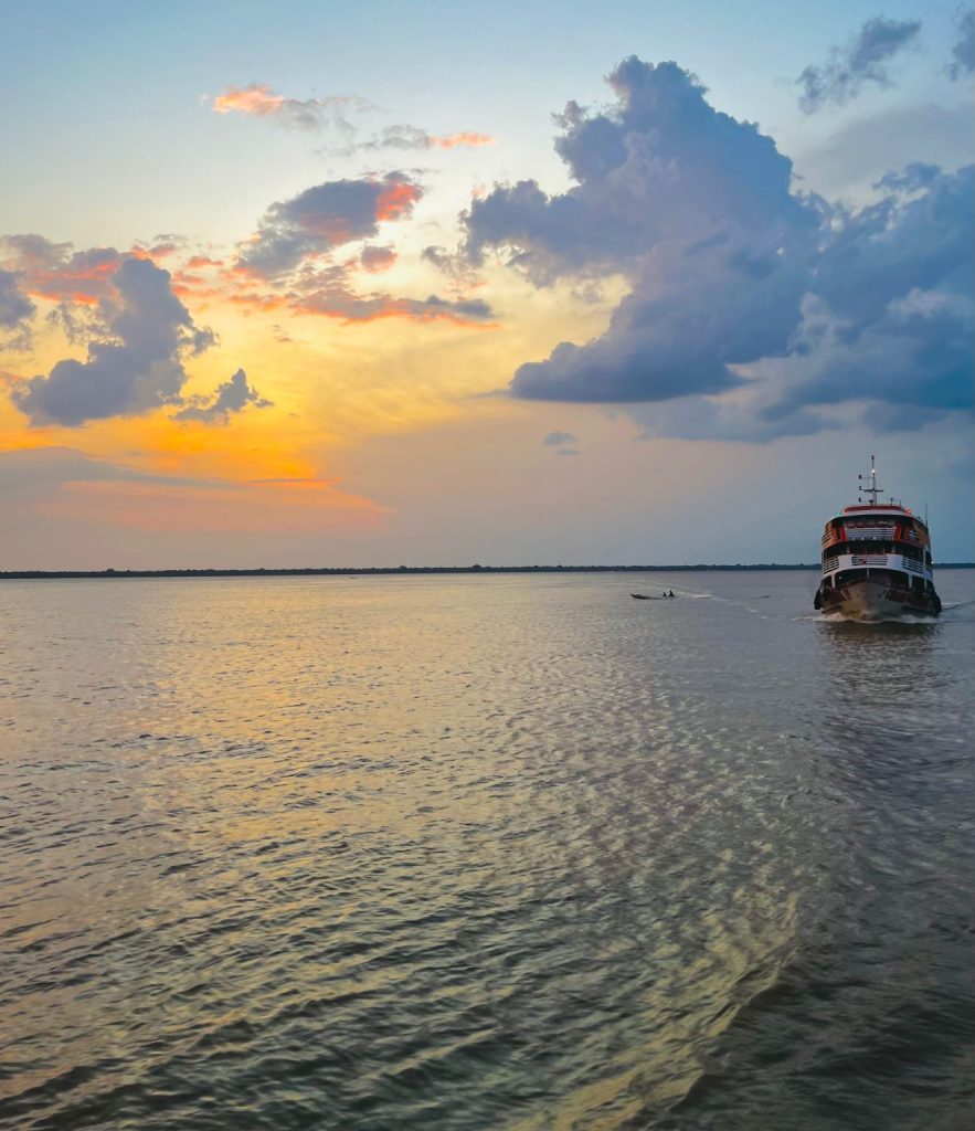 Barco estilo catamarã navegando sobre um rio bastante largo e o sol se pondo ao fundo.